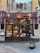 A charming storefront of an Italian shop named 'La Prosciutteria' with rustic wooden doors and a sign displaying various specialties such as 'panini' and 'merende.' The facade is adorned with vibrant purple flowers, adding a touch of elegance. Inside, shelves are filled with food products, creating a warm and inviting atmosphere.