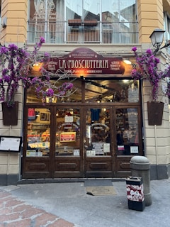 A charming storefront of an Italian shop named 'La Prosciutteria' with rustic wooden doors and a sign displaying various specialties such as 'panini' and 'merende.' The facade is adorned with vibrant purple flowers, adding a touch of elegance. Inside, shelves are filled with food products, creating a warm and inviting atmosphere.