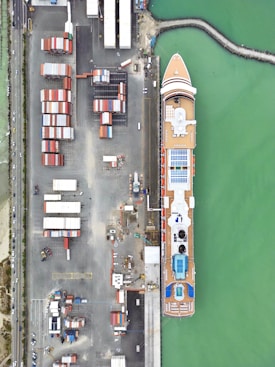 Aerial view of a large cruise ship docked at a busy port. Numerous colorful shipping containers are neatly arranged in stacks on the dock nearby. The ship appears well-ordered with visible blue swimming pools and orange decks. The surrounding water is a vibrant green, and there are various small vehicles and equipment scattered around the port area.
