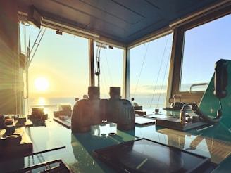 A ship's control room with a wide view of the ocean during sunset. The sunlight floods the room, illuminating various navigation instruments and equipment, including binoculars and communication devices, all set against a reflective surface.