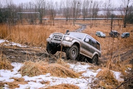A large SUV is navigating a muddy off-road terrain, with other similar vehicles visible in the background. The landscape is characterized by dry grass and patches of snow, while the sky looks overcast. Leafless trees surround the area, adding to the stark and rugged environment.