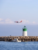 A commercial airplane with orange and white livery flies low above a calm seascape. In the foreground, a small motorboat with a person onboard is near a rocky breakwater. A green and white lighthouse stands on the rocks, and the scene is set under a clear blue sky with scattered clouds.