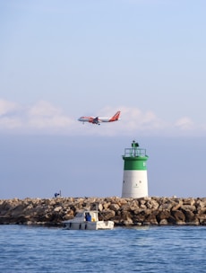 A commercial airplane with orange and white livery flies low above a calm seascape. In the foreground, a small motorboat with a person onboard is near a rocky breakwater. A green and white lighthouse stands on the rocks, and the scene is set under a clear blue sky with scattered clouds.