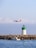 A commercial airplane with orange and white livery flies low above a calm seascape. In the foreground, a small motorboat with a person onboard is near a rocky breakwater. A green and white lighthouse stands on the rocks, and the scene is set under a clear blue sky with scattered clouds.
