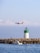 A commercial airplane with orange and white livery flies low above a calm seascape. In the foreground, a small motorboat with a person onboard is near a rocky breakwater. A green and white lighthouse stands on the rocks, and the scene is set under a clear blue sky with scattered clouds.