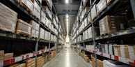 A large warehouse aisle flanked by tall metal shelves filled with stacked boxes and packages. The shelves extend towards the back of the warehouse, where they converge in the distance. The lighting is bright and industrial, with fluorescent lights illuminating the space.