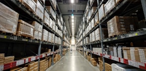A large warehouse aisle flanked by tall metal shelves filled with stacked boxes and packages. The shelves extend towards the back of the warehouse, where they converge in the distance. The lighting is bright and industrial, with fluorescent lights illuminating the space.