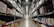 A large warehouse aisle flanked by tall metal shelves filled with stacked boxes and packages. The shelves extend towards the back of the warehouse, where they converge in the distance. The lighting is bright and industrial, with fluorescent lights illuminating the space.