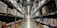 A large warehouse aisle flanked by tall metal shelves filled with stacked boxes and packages. The shelves extend towards the back of the warehouse, where they converge in the distance. The lighting is bright and industrial, with fluorescent lights illuminating the space.