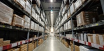 A large warehouse aisle flanked by tall metal shelves filled with stacked boxes and packages. The shelves extend towards the back of the warehouse, where they converge in the distance. The lighting is bright and industrial, with fluorescent lights illuminating the space.