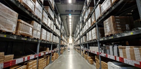 A large warehouse aisle flanked by tall metal shelves filled with stacked boxes and packages. The shelves extend towards the back of the warehouse, where they converge in the distance. The lighting is bright and industrial, with fluorescent lights illuminating the space.
