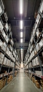A vast warehouse aisle lined with tall shelves stacked with numerous large boxes. The perspective is down the center of the aisle, with fluorescent lights illuminating the space overhead.