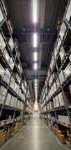 A vast warehouse aisle lined with tall shelves stacked with numerous large boxes. The perspective is down the center of the aisle, with fluorescent lights illuminating the space overhead.