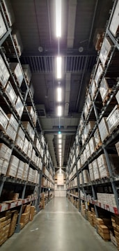 A vast warehouse aisle lined with tall shelves stacked with numerous large boxes. The perspective is down the center of the aisle, with fluorescent lights illuminating the space overhead.