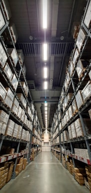 A vast warehouse aisle lined with tall shelves stacked with numerous large boxes. The perspective is down the center of the aisle, with fluorescent lights illuminating the space overhead.