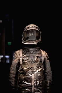 A vintage space suit is displayed against a dark background. The suit appears metallic with a reflective surface and intricate details like straps and fasteners.