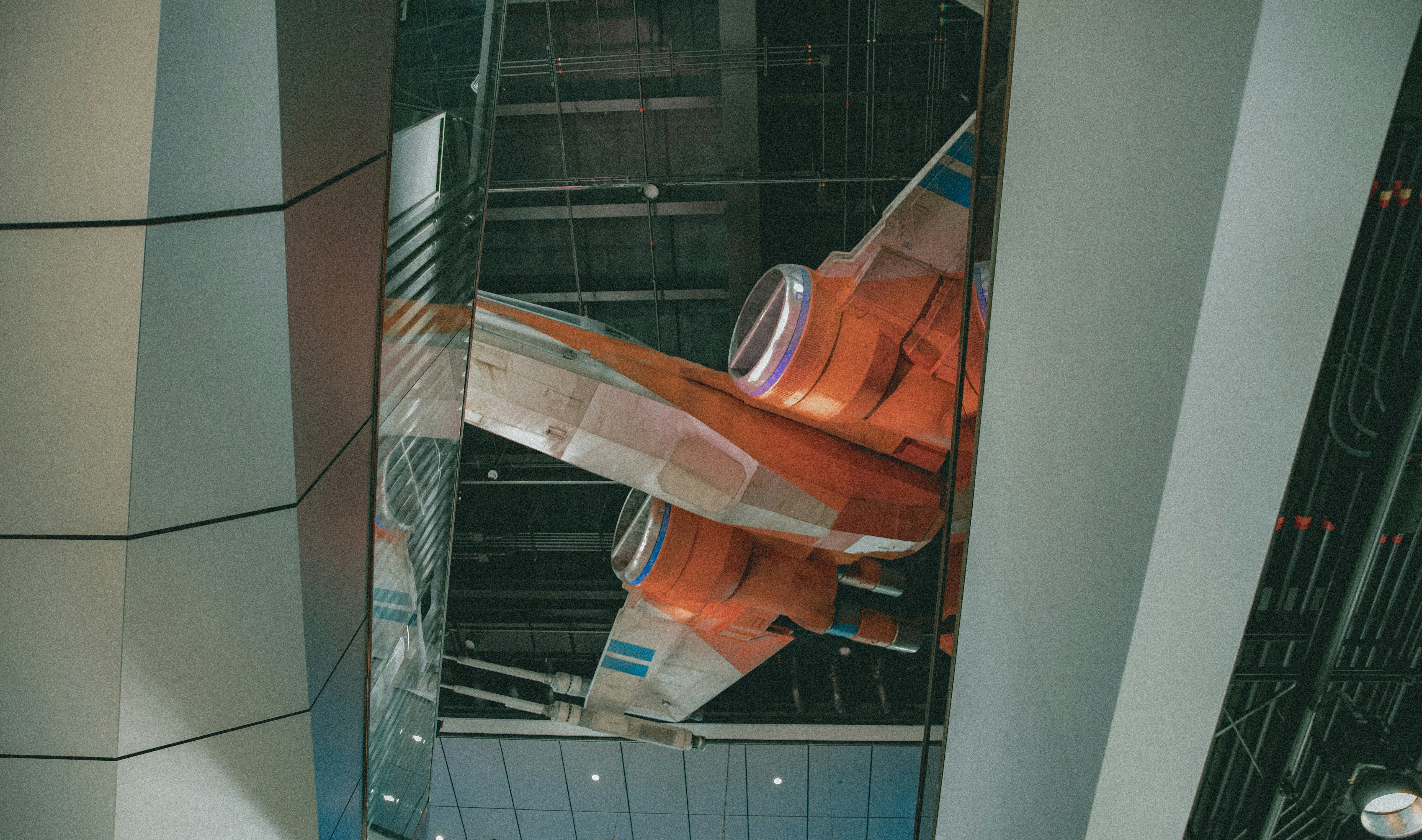 A futuristic orange and white spacecraft is displayed within an indoor setting that features a modern architectural design. The craft appears suspended with visible engines and sleek lines, highlighted by overhead lighting. The surrounding environment consists of large metallic and glass panels.