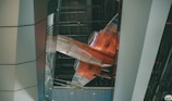A futuristic orange and white spacecraft is displayed within an indoor setting that features a modern architectural design. The craft appears suspended with visible engines and sleek lines, highlighted by overhead lighting. The surrounding environment consists of large metallic and glass panels.