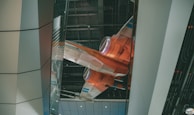 A futuristic orange and white spacecraft is displayed within an indoor setting that features a modern architectural design. The craft appears suspended with visible engines and sleek lines, highlighted by overhead lighting. The surrounding environment consists of large metallic and glass panels.