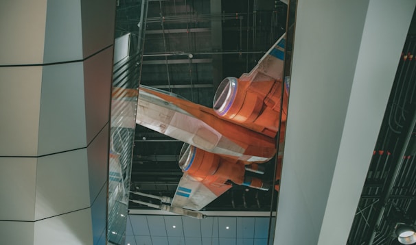 A futuristic orange and white spacecraft is displayed within an indoor setting that features a modern architectural design. The craft appears suspended with visible engines and sleek lines, highlighted by overhead lighting. The surrounding environment consists of large metallic and glass panels.