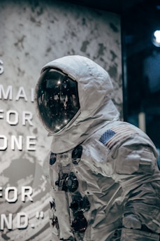 A display of a space suit with a reflective visor is showcased. The suit is adorned with various patches, including the United States flag, and features multiple control modules on the chest. The background includes a textured wall with partially visible text, creating an exhibition-like setting.
