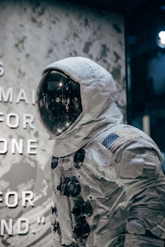 A display of a space suit with a reflective visor is showcased. The suit is adorned with various patches, including the United States flag, and features multiple control modules on the chest. The background includes a textured wall with partially visible text, creating an exhibition-like setting.