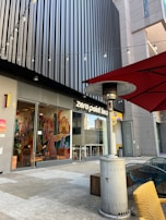 Outdoor seating area of a modern tea shop with colorful wall art visible through large glass windows. There are string lights overhead, a tall outdoor heater, and a red parasol with yellow and blue chairs in view. The building features a sleek, vertical panel facade.