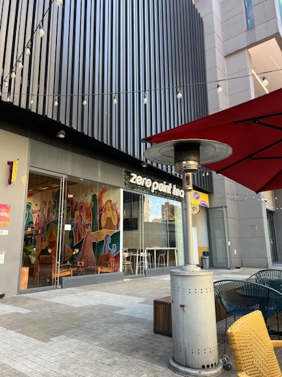 Outdoor seating area of a modern tea shop with colorful wall art visible through large glass windows. There are string lights overhead, a tall outdoor heater, and a red parasol with yellow and blue chairs in view. The building features a sleek, vertical panel facade.
