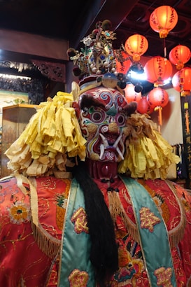 An intricately detailed, colorful traditional mask with elaborate patterns and vibrant hues. The mask is topped with a decorative headdress featuring various ornaments, and the shoulders are adorned with large, ruffled yellow decorations. In the background, red lanterns hang from the ceiling, adding to the festive and cultural atmosphere.