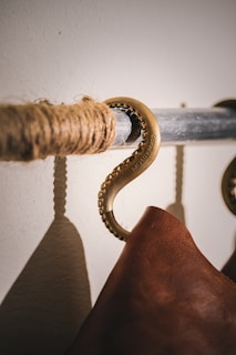 A close-up of a brass hook attached to a metal rod wrapped with rope. A leather item hangs from the hook, casting a shadow on the textured wall, highlighting the craftsmanship of the materials.