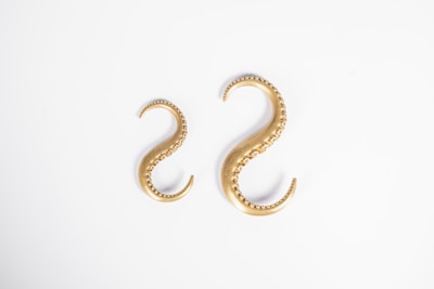 Two gold-colored, S-shaped objects resembling octopus tentacles with textured details and smooth curves placed against a white background.