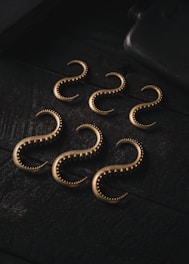 A set of six metallic, curved hooks with a textured surface are arranged on a dark, textured surface. The hooks have an antique bronze appearance and are uniformly spaced, creating a visually interesting pattern.