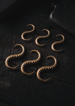 A set of six metallic, curved hooks with a textured surface are arranged on a dark, textured surface. The hooks have an antique bronze appearance and are uniformly spaced, creating a visually interesting pattern.