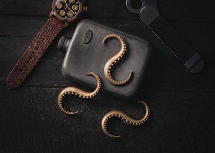 A composition with a dark metallic flask engraved with '1914', surrounded by brass octopus tentacle-shaped objects with 'COPPERTIST.WU' inscribed on them. A leather-strapped watch with a black and gold face, alongside a darkly toned metallic tool, complements the arrangement. The textures of leather, metal, and the unique shape of the tentacles add interest.