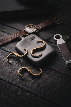 A metallic flask labeled with '1914' is placed on a wooden surface. Gold-colored tentacle-like accessories surround the flask. A vintage watch with a brown leather strap, a metal bottle opener, and a portion of a closed book are also arranged around the flask. The lighting is dramatic, emphasizing the shine of the metals and the texture of the wood.