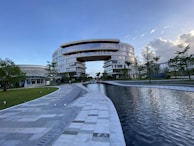 A large, modern building with a unique arch design dominates the scene, surrounded by lush greenery and a reflective water feature in the foreground. The architecture is characterized by smooth, curved lines and extensive use of glass, providing a futuristic and sophisticated appearance. Clear skies with some clouds contribute to a serene atmosphere.