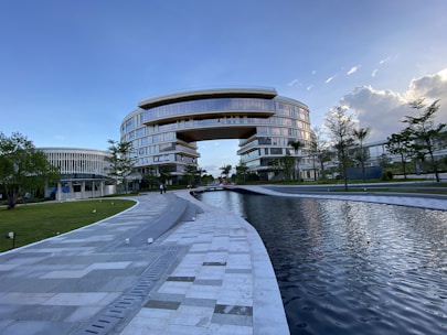 A large, modern building with a unique arch design dominates the scene, surrounded by lush greenery and a reflective water feature in the foreground. The architecture is characterized by smooth, curved lines and extensive use of glass, providing a futuristic and sophisticated appearance. Clear skies with some clouds contribute to a serene atmosphere.