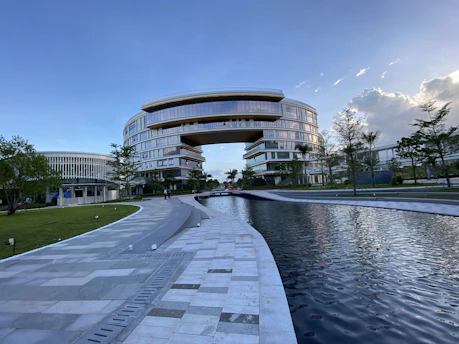 A large, modern building with a unique arch design dominates the scene, surrounded by lush greenery and a reflective water feature in the foreground. The architecture is characterized by smooth, curved lines and extensive use of glass, providing a futuristic and sophisticated appearance. Clear skies with some clouds contribute to a serene atmosphere.
