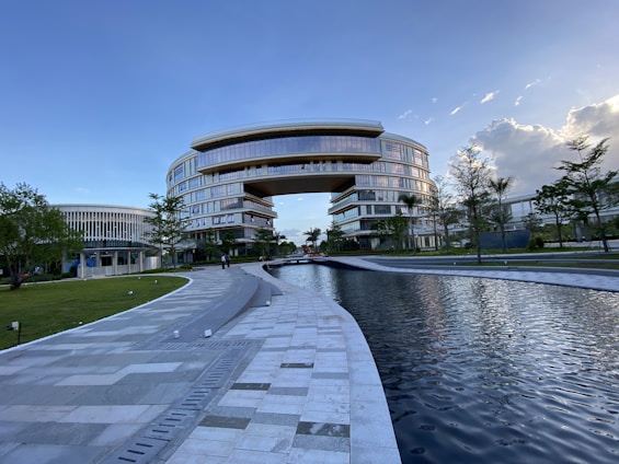A large, modern building with a unique arch design dominates the scene, surrounded by lush greenery and a reflective water feature in the foreground. The architecture is characterized by smooth, curved lines and extensive use of glass, providing a futuristic and sophisticated appearance. Clear skies with some clouds contribute to a serene atmosphere.