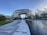 A large, modern building with a unique arch design dominates the scene, surrounded by lush greenery and a reflective water feature in the foreground. The architecture is characterized by smooth, curved lines and extensive use of glass, providing a futuristic and sophisticated appearance. Clear skies with some clouds contribute to a serene atmosphere.