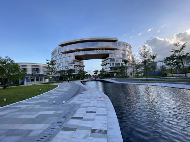 A large, modern building with a unique arch design dominates the scene, surrounded by lush greenery and a reflective water feature in the foreground. The architecture is characterized by smooth, curved lines and extensive use of glass, providing a futuristic and sophisticated appearance. Clear skies with some clouds contribute to a serene atmosphere.