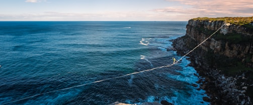 A daring adventurer navigates a highline stretched across a vast seascape from a rocky cliff. The ocean spreads out below, meeting the rugged coastline with waves crashing against the rocks. The clear blue sky is tinged with soft clouds on the horizon.