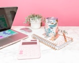 A neatly arranged desk setup includes a laptop with colorful keyboard covers, a small potted plant, a floral-patterned tumbler, a spiral-bound notebook with geometric design and positive message, a pink calculator, and a golden pen.