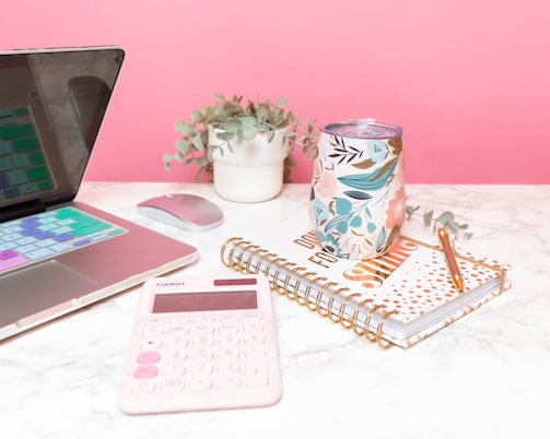 A neatly arranged desk setup includes a laptop with colorful keyboard covers, a small potted plant, a floral-patterned tumbler, a spiral-bound notebook with geometric design and positive message, a pink calculator, and a golden pen.