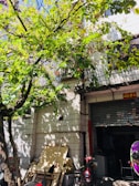 A tree with lush green leaves casts shadows on a white tiled building. Various objects, including a broom and wooden boards, are stacked against the building. A red bike is parked next to a small garage-like entrance with a partially open shutter. There is also a vibrant purple and green element in the foreground.