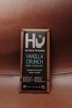 A chocolate bar wrapped in brown packaging with a focus on a square label featuring the brand name Hu and the message 'Get Back to Human.' The product is labeled as Vanilla Crunch Dark Chocolate with 70% organic cacao, vegan, primal, and free from refined sugars, dairy, soy, and other additives.