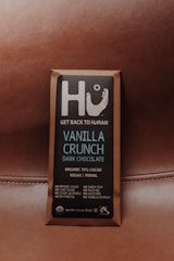 A chocolate bar wrapped in brown packaging with a focus on a square label featuring the brand name Hu and the message 'Get Back to Human.' The product is labeled as Vanilla Crunch Dark Chocolate with 70% organic cacao, vegan, primal, and free from refined sugars, dairy, soy, and other additives.