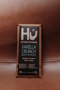 A chocolate bar wrapped in brown packaging with a focus on a square label featuring the brand name Hu and the message 'Get Back to Human.' The product is labeled as Vanilla Crunch Dark Chocolate with 70% organic cacao, vegan, primal, and free from refined sugars, dairy, soy, and other additives.