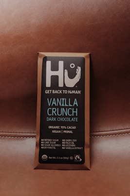 A chocolate bar wrapped in brown packaging with a focus on a square label featuring the brand name Hu and the message 'Get Back to Human.' The product is labeled as Vanilla Crunch Dark Chocolate with 70% organic cacao, vegan, primal, and free from refined sugars, dairy, soy, and other additives.