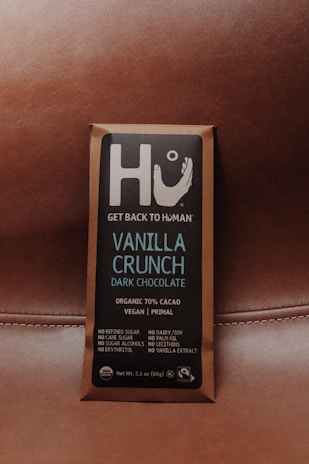 A chocolate bar wrapped in brown packaging with a focus on a square label featuring the brand name Hu and the message 'Get Back to Human.' The product is labeled as Vanilla Crunch Dark Chocolate with 70% organic cacao, vegan, primal, and free from refined sugars, dairy, soy, and other additives.